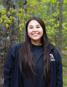 Cheyanne is looking and smiling at the camera. They have long black hair and standing in front of leafy green trees.