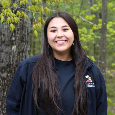 Cheyanne is looking and smiling at the camera. They have long black hair and standing in front of leafy green trees.